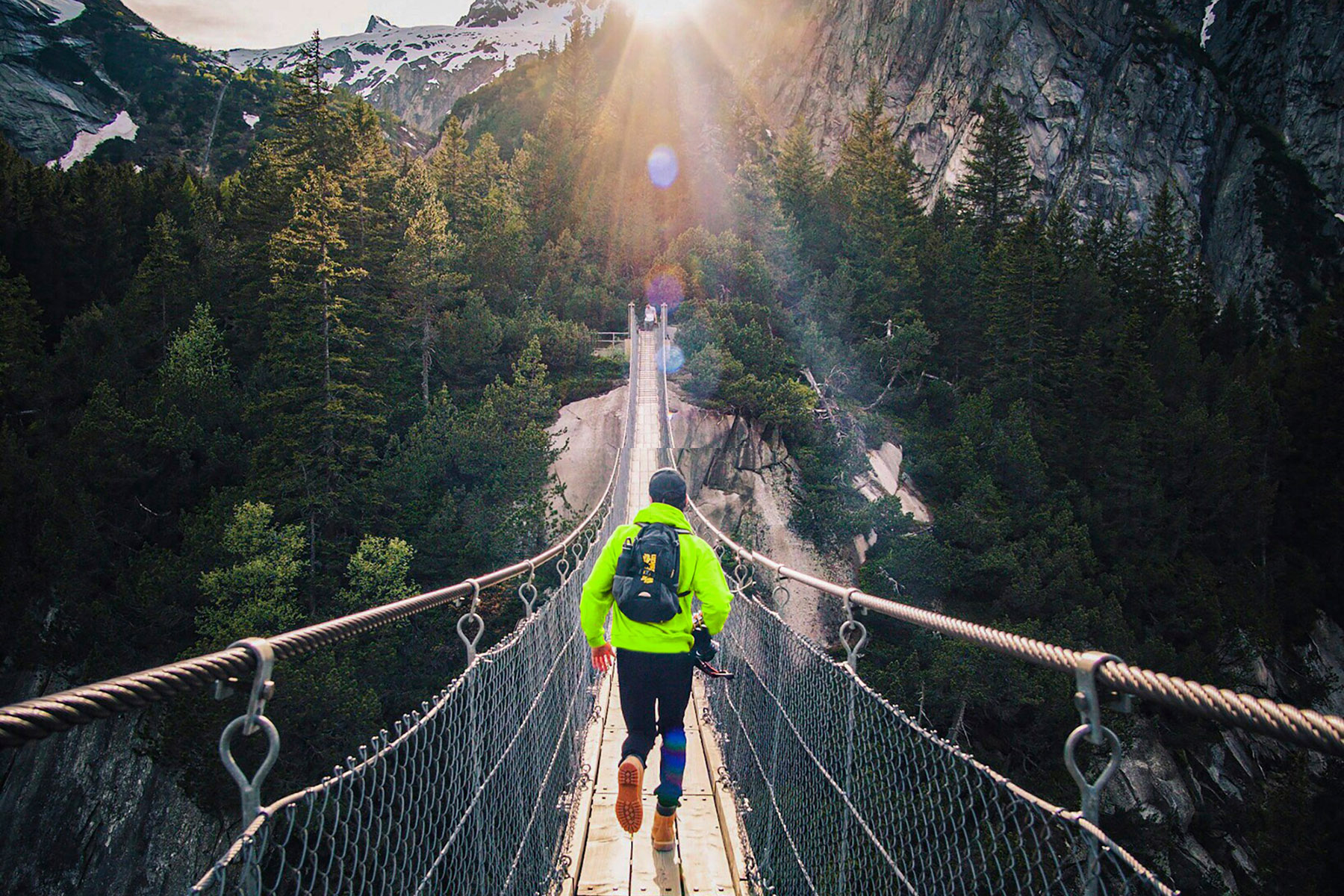 Mann in neoongelben Oberteil und Rucksack laufend auf Hängebrücke vor Bergkulisse