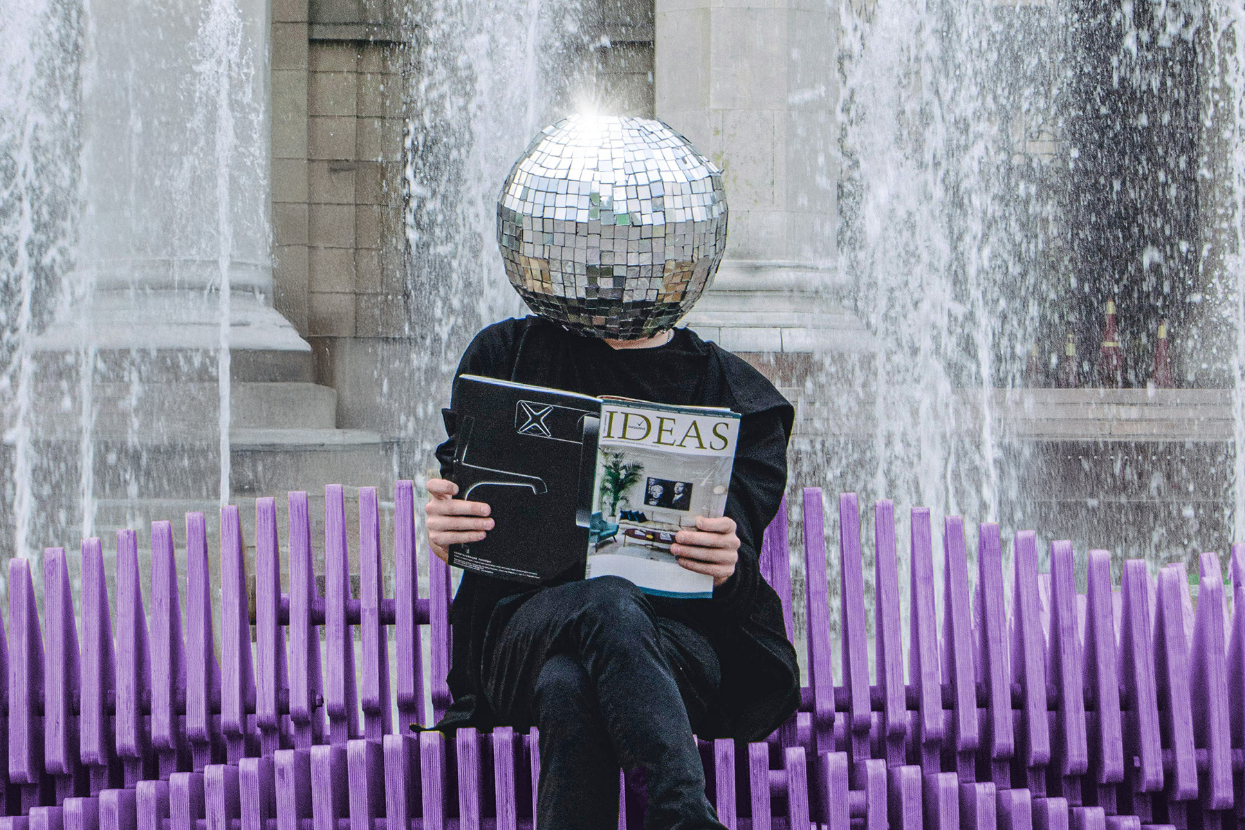 Person sitzt auf einer lila Bank mit Zeitung in der Hand und Glitzerkugel auf dem Kopf